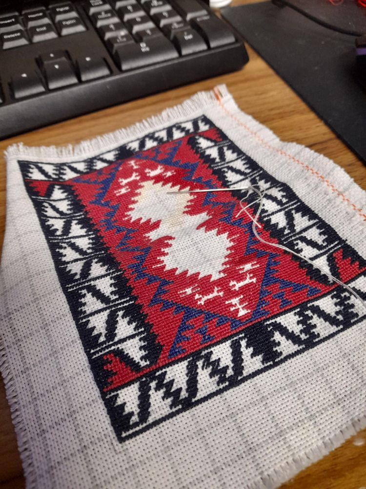 A tiny needlepoint embriodery being completed sitting on a wooden desk. It is surrounded by a keyboard above it and a mouse pad with a mouse on its right. 