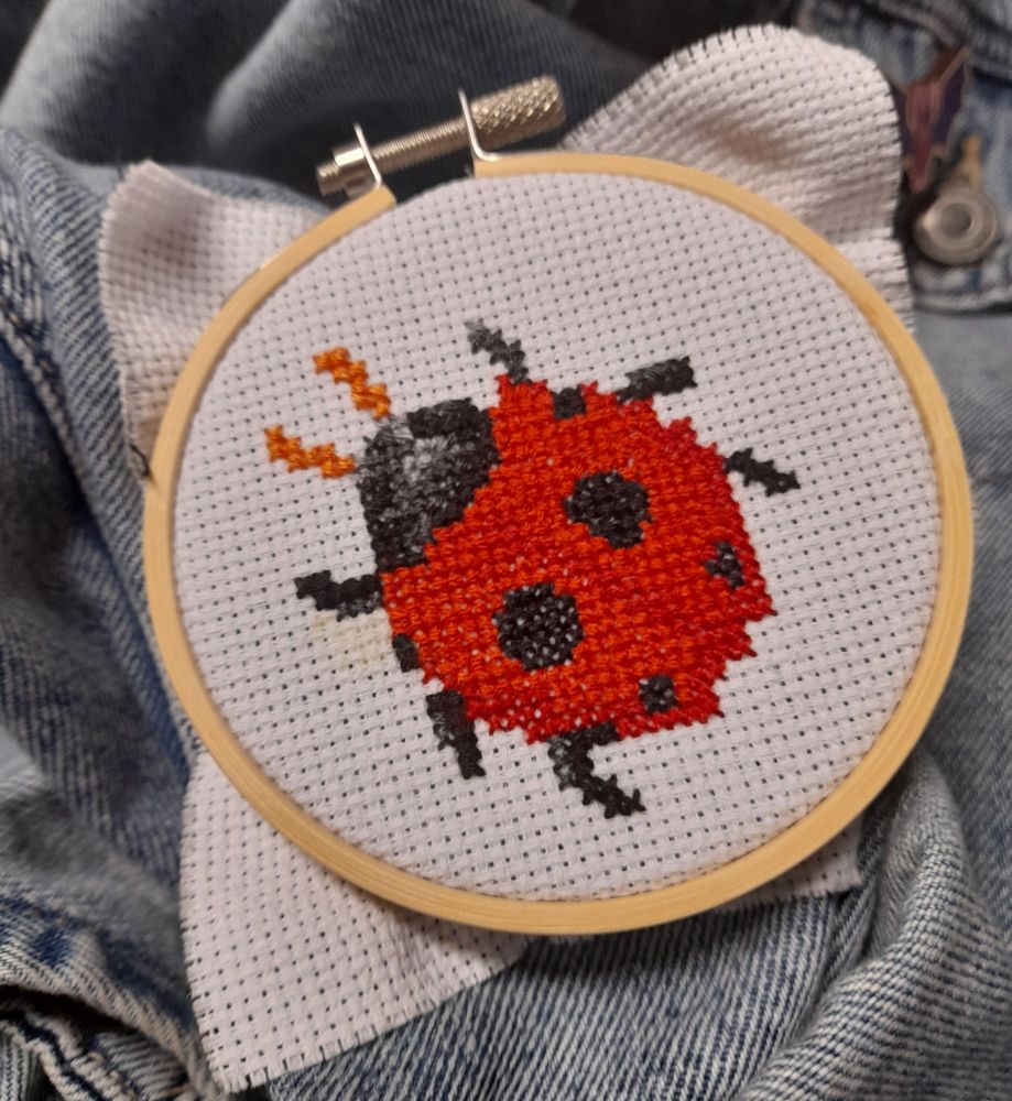 A finished ladybug cross stitch sitting in an embroidery hoop on a denim jacket. 