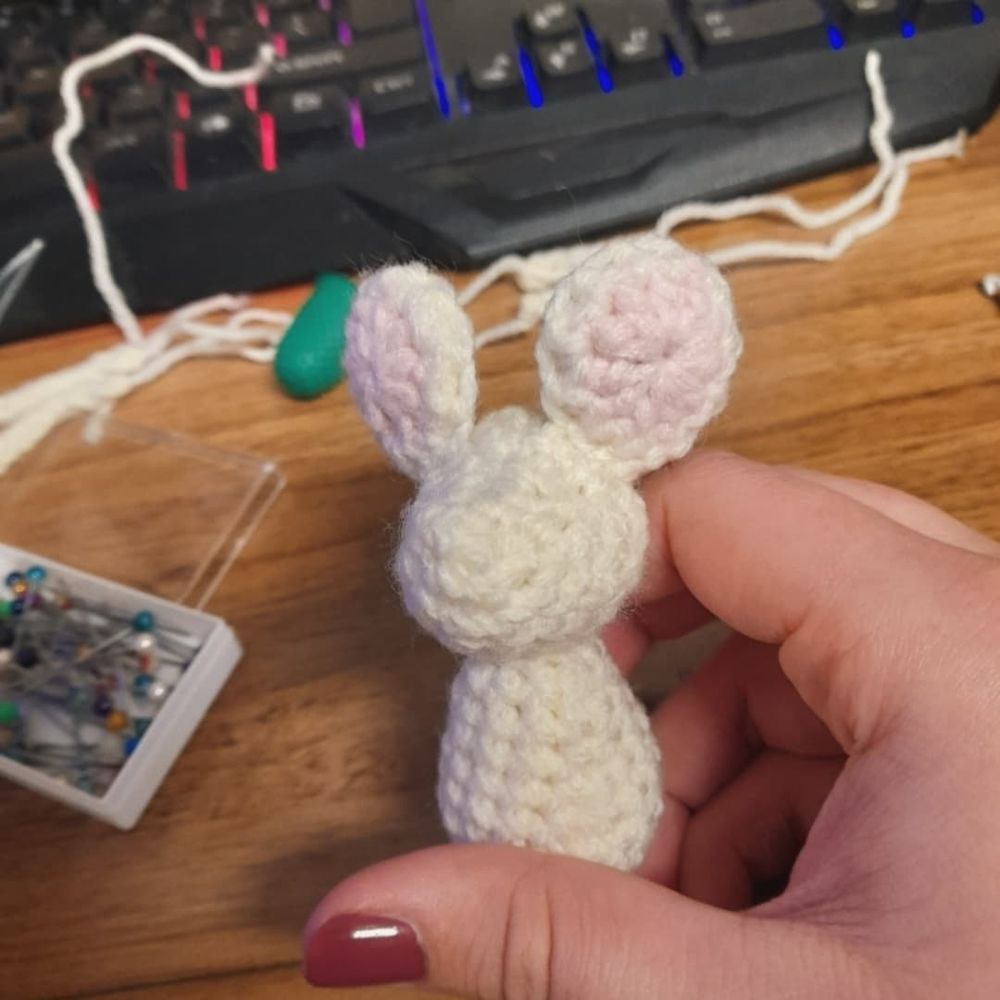 A half-finished mouse is being held, having only a body, head, and ears. In the background a thimble and a box of pins can be seen resting on a wooden desk.