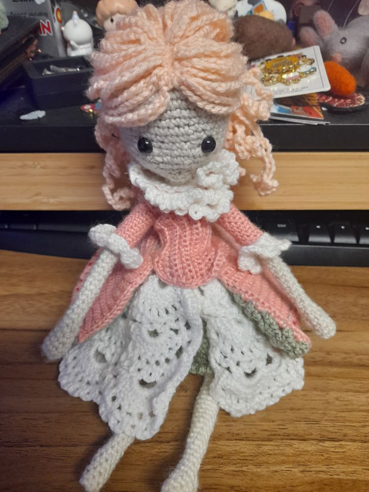 A crochet doll sits on a wooden desk, almost finished. She leans against a tucked away keyboard and a desk drawer, miscellaneous trinkets and clutter are behind her. She has button eyes but no smile or blush on her face. Her pink hair is tied back, and her clothes have been finished. She is wearing a dark pink blouse on top of a Layered skirt. She has no shoes on. 