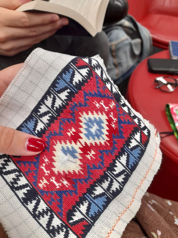 A tiny needlepoint embriodery being held by a feminine hand with red bail polish. In the background is person reading. 