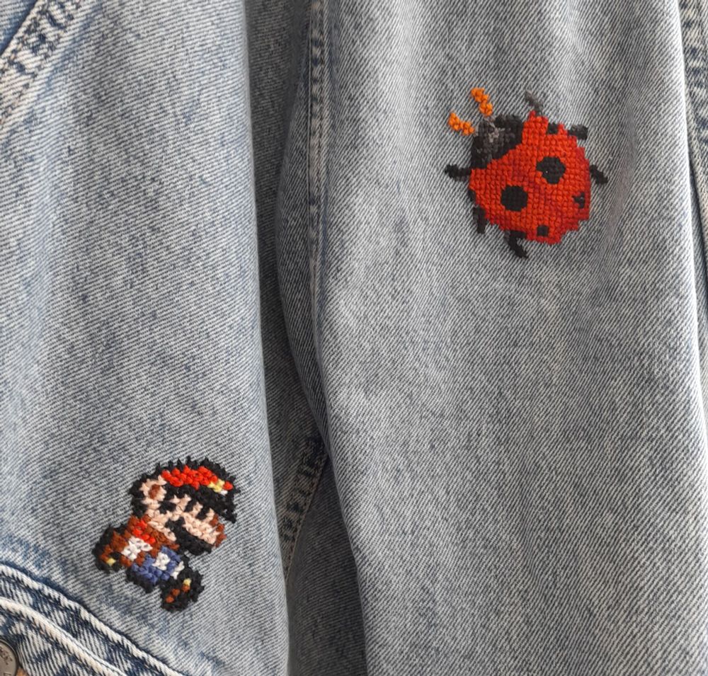 Two tint cross stitch patterns on a denim jacket. One in the style of paper mario as he walks along the hem, and the other is a red ladybug on the jackets sleeve. 