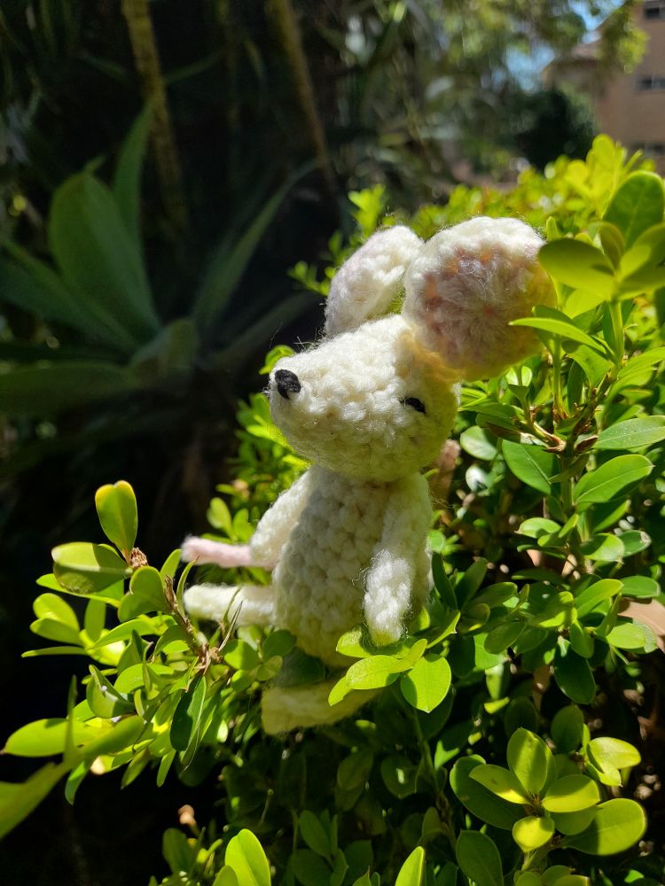 Crochet mouse sitting with closed eyes on a hedge. There are large green plants in the background. 