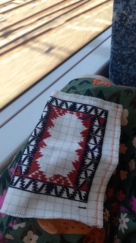 A tiny needlepoint embriodery that is only in its beginning stages. It is rested on the lap on someone on the train wearing a green floral dress. It is next to a train window, where blurry train tracks can be seen in the background  