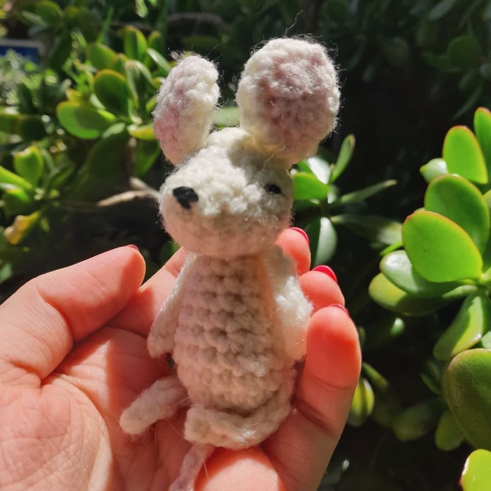 Small crochet mouse sits in a feminine hand that has red painted nails. In the background are various plants. 