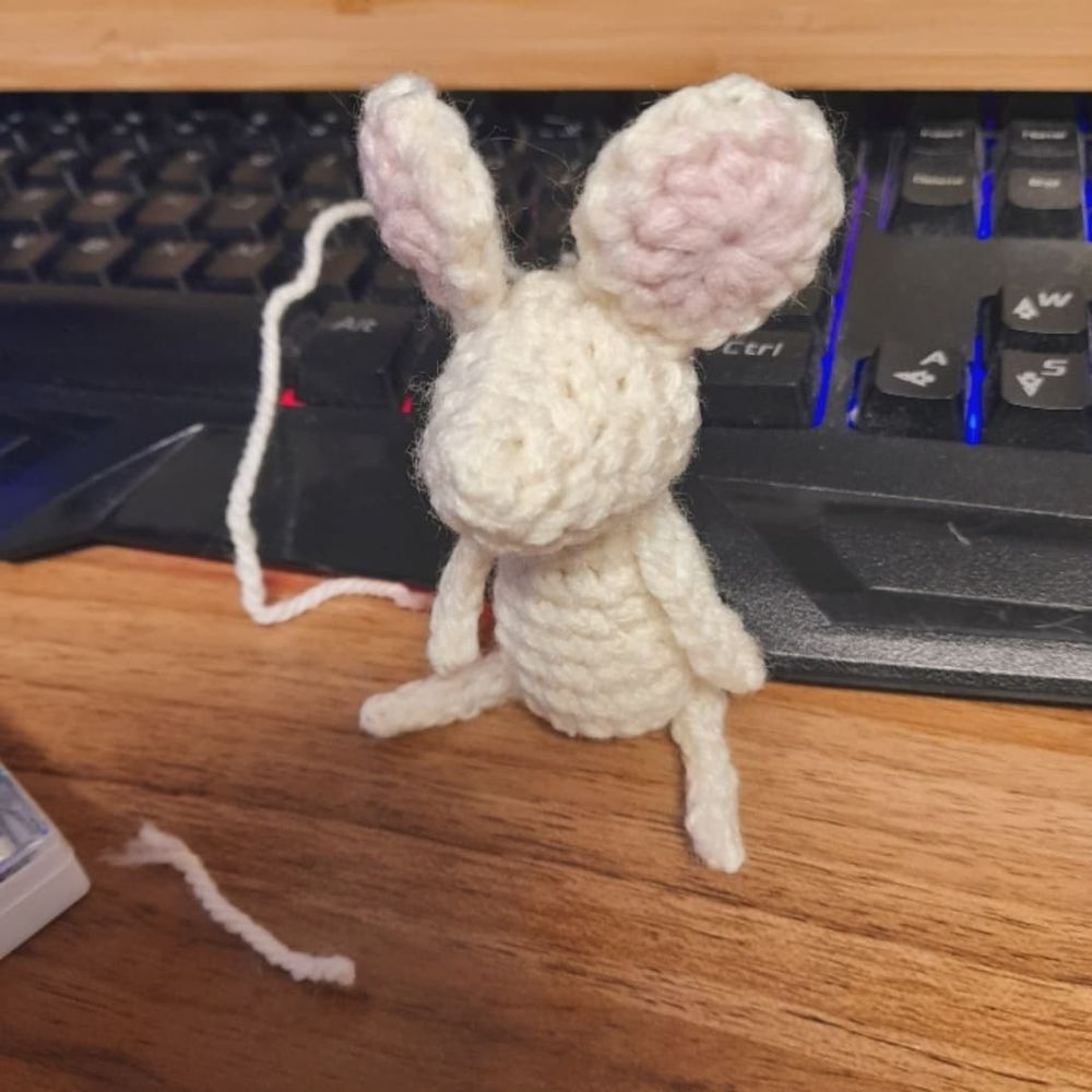 A half-finished crochet mouse sits against a keyboard on a wooden desk.