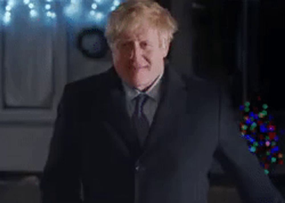 a man in a suit and tie is standing in front of a christmas tree and giving a thumbs up .