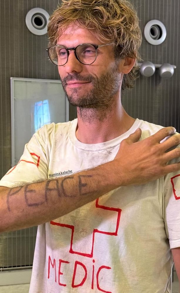 Photo of Dr. Baptiste André on arrival in Paris, in a t-shirt with a red cross  that says "MEDIC", and with his arm showing the word "PEACE". 