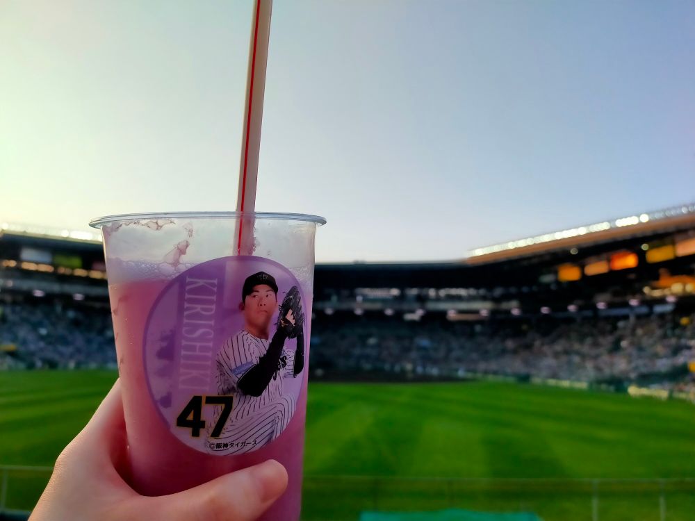 プラスチックカップに入ったぶどうジュースを持っている。後ろに甲子園球場のグラウンドが写っている