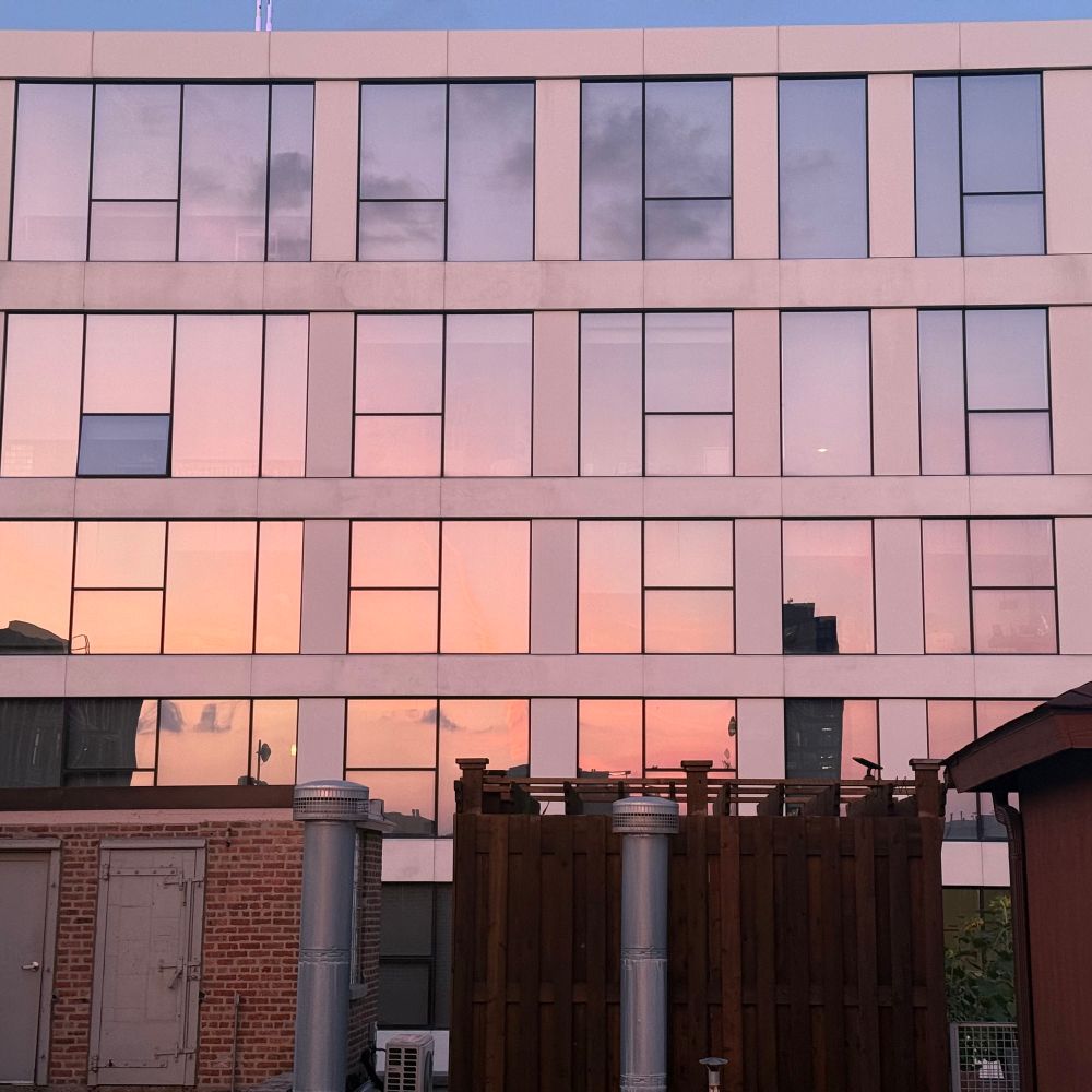 The orange purple sky at sunset reflected in the glass of an apartment with some rooftop structures in the foreground.