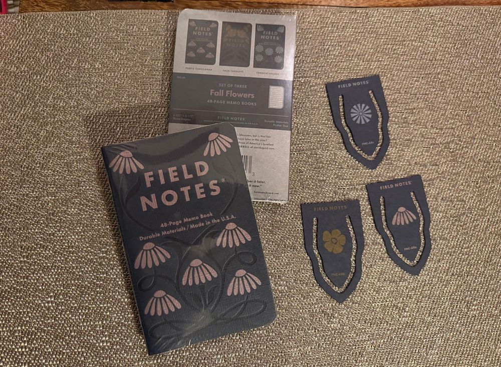 The still-wrapped front and back covers of a 3-pack of Field Notes notebooks with embossed and textured covers depicting fall season wildflowers. There are also three bookmarks, each depicting one of the three wildflowers on the covers of the notebooks.
