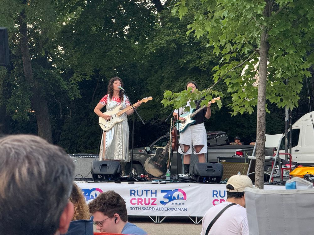 The band Finom onstage performing at Kilbourn Park in Chicago with Sima Cunningham (left) and Macie Stewart (right, obscured by a tree branch)