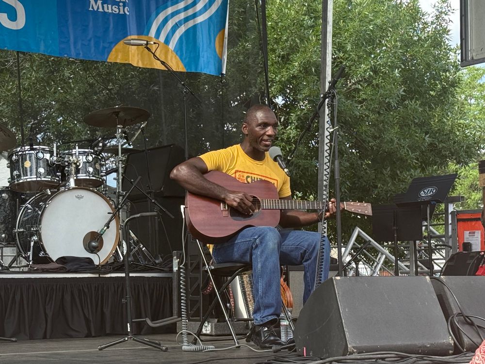Cedric Burnside seated onstage at Square Roots Festival playing acoustic guitar 