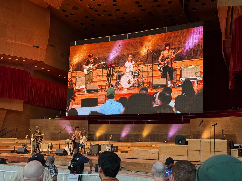 The band Horsegirl onstage at Pritzker Pavilion playing in front of a jumbo screen of them performing 