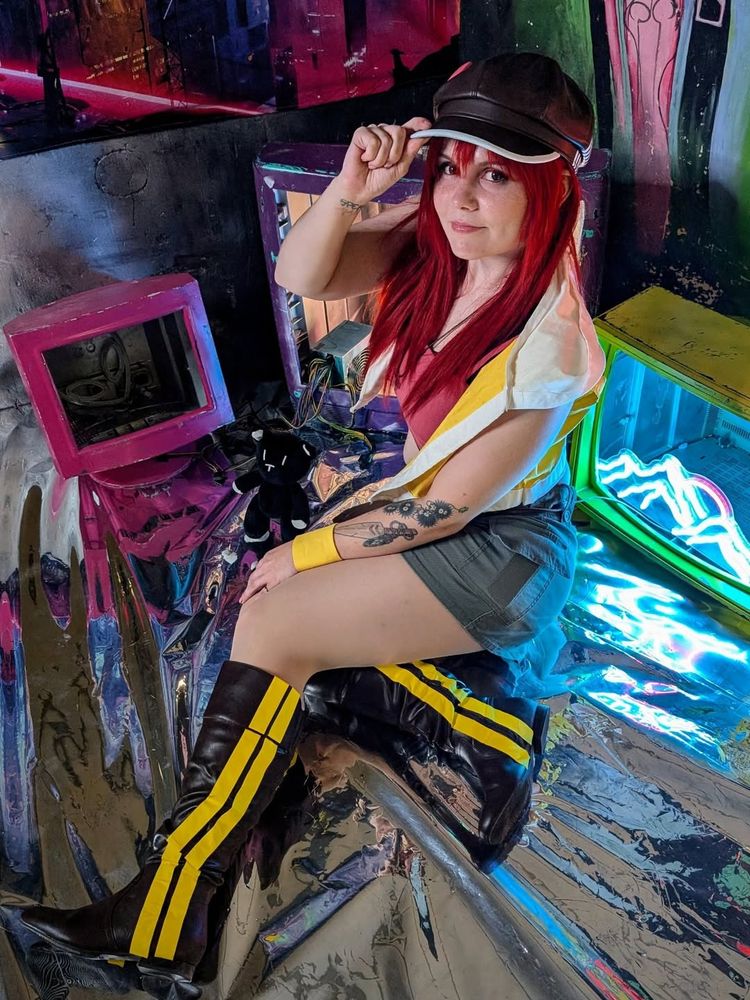 A cosplayer dressed as Shiki Misaki from The World Ends With You poses in a colourful setting with neon lights and retro TVs. She is seated on a reflective silver surface, lifting her hat with one hand and smiling slightly at the camera. A small plush Mr. Mew stands beside her.