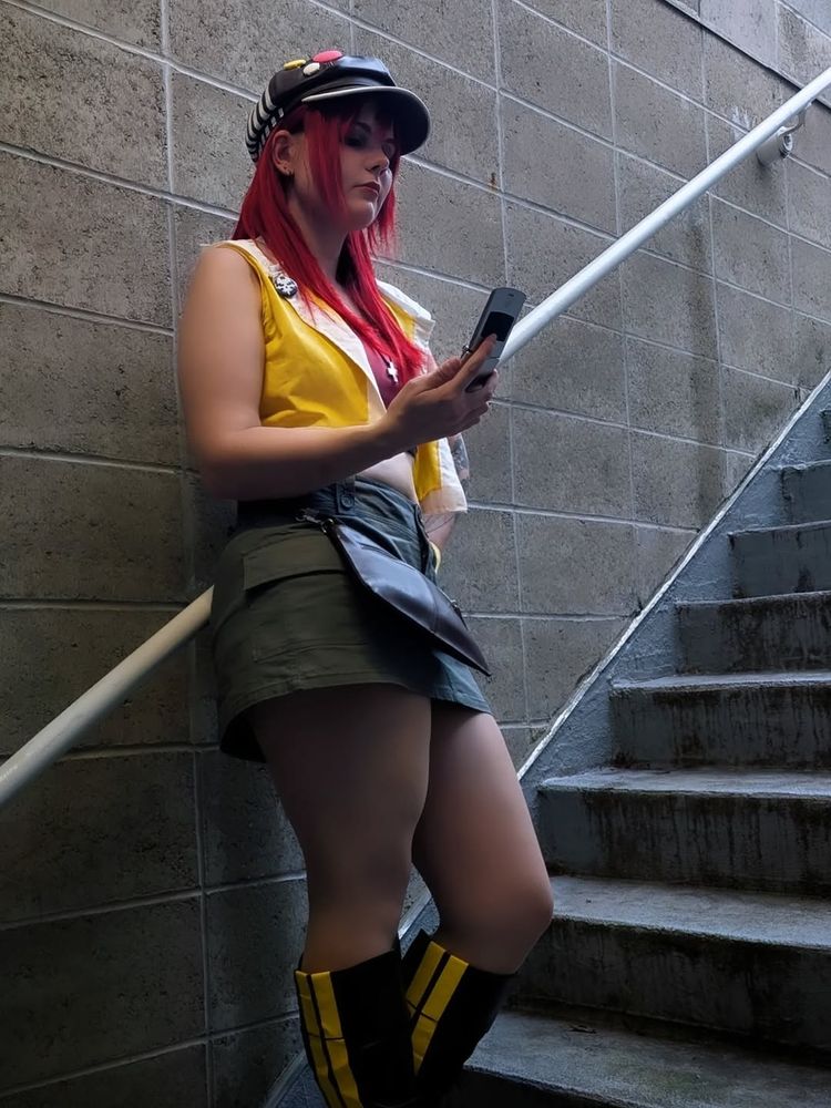 A person in a Shiki Misaki cosplay from The World Ends With You stands on a concrete stairwell, leaning casually against the railing while looking at a flip phone.