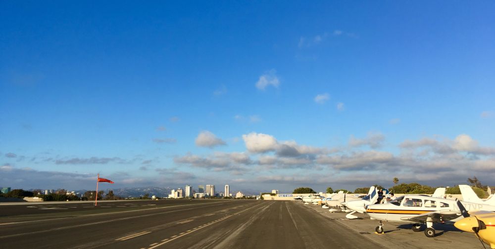 Picture looking up a runway, small airplanes to the right, buildings in the distance. Clear blue skies overhead with just a few small clouds farther away off the airport.