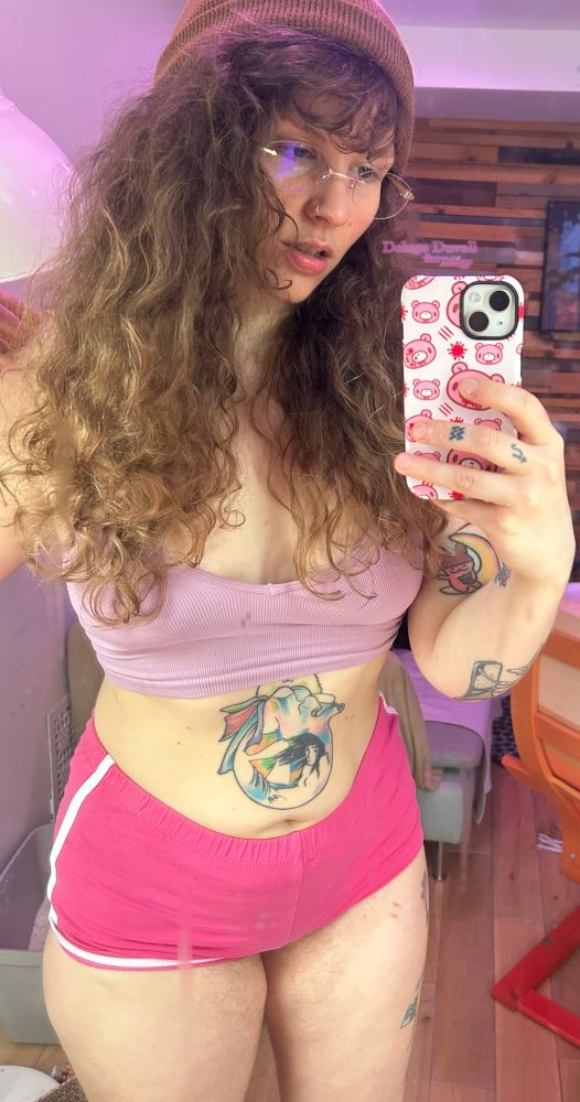 Deluge takes a mirror selfie wearing a brown beanie, pair of frameless glasses, a pink crop top, and pink booty shorts. Her long curly hair is down.