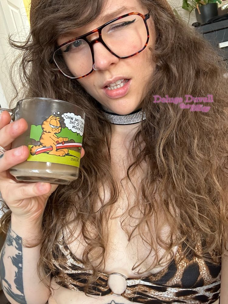 Deluge takes a selfie wearing brown tortoishell glasses, a brown beanie, and a leopard print halter crop top. Their long curly hair is down and they are winking at the camera while holding up vintage mcdonald’s garfield the cat mug full of coffee, which are known to contain lead.