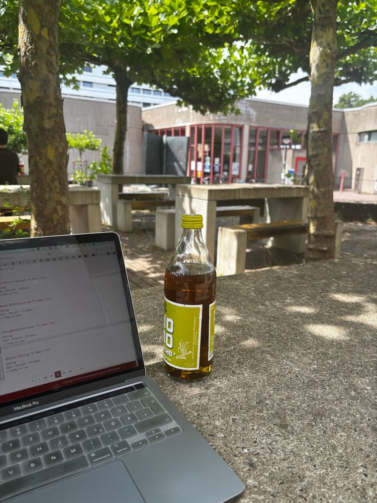 Laptop und Mate stehen draussen auf einem Betontisch. Im Hintergrund weitere Betontische mit Sitzflächen unter grünen Weiden, dahinter universitärer kahler Betonbau und eine Heine-Skulptur von Bert Gerresheim