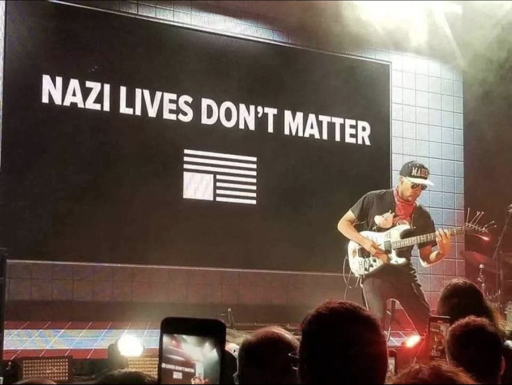 Tom Morello do Rage Against the Machine tocando guitarra no palco com uma bandeira preta com letras brancas ao fundo escrito: "Vidas nazistas não importam."