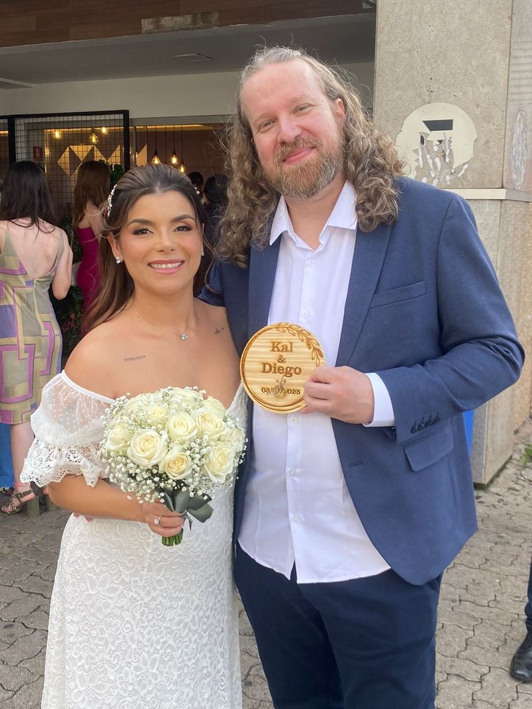 A noiva e o noivo posando na frente do cartório, a noiva segurando um buquê de rosas brancas e o noivo uma placa de madeira gravado Kal e Diego 03/01/25 e com as alianças amarradas nela