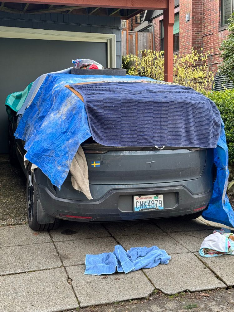 A newer car is parked in the driveway of a house. It’s top is covered in layers of blue plastic tarps, floor mats, and bath towels