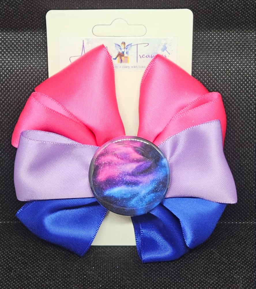 A hand sewn hair bow layered in bisexual flag colors, Pink, Purple and Blue. In the center is a circular gem with a galaxy image on it in the same pink, purple and blue.