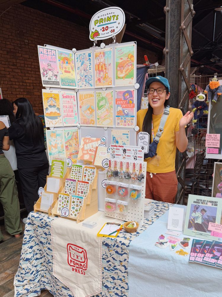 Half table setup with riso prints, sticker sheets, notepads, charms, and patches 