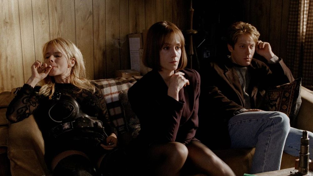 screenshot from 'crash' (1996), showing james spader and holly hunter sitting on a couch attentively watching the tv while rosanna arquette sits next to them smoking