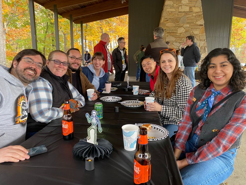 Picture taken looking down a picnic table, where 7 people smile for the camera. Given how they’re dressed it looks chilly. 