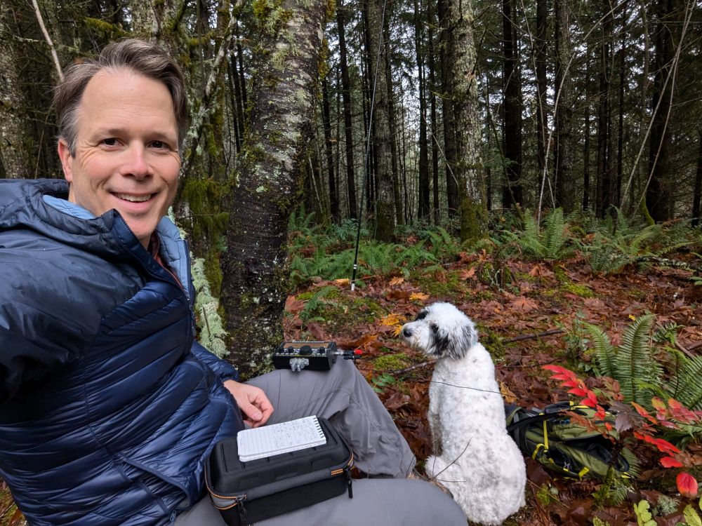 I am outdoors in a forested area, sitting on the ground and smiling at the camera. I am wearing a puffy blue jacket and have some equipment in front of them, including a notebook and a small case with, with an Elecraft KX2 on my left knee. Next to me is a fluffy white and black dog is sitting, looking off into the distance. The background is lush with mossy trees, ferns, and fallen leaves, creating a peaceful, natural setting.