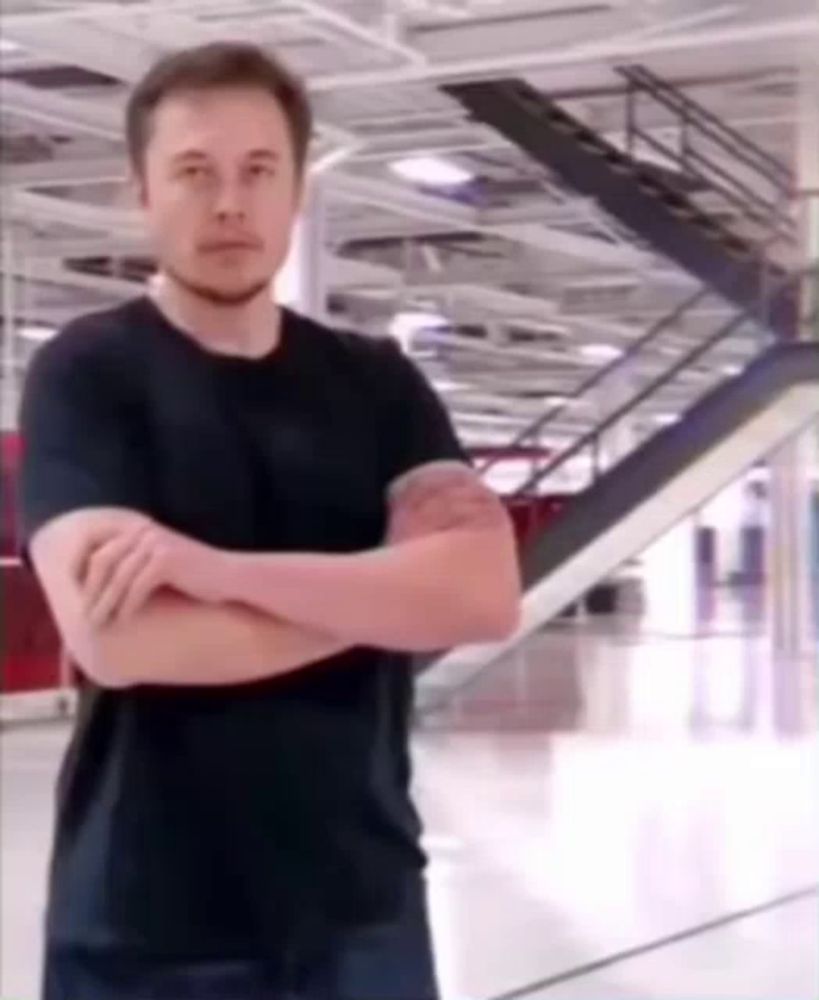 elon musk is standing in a room with his arms crossed and looking at the camera .