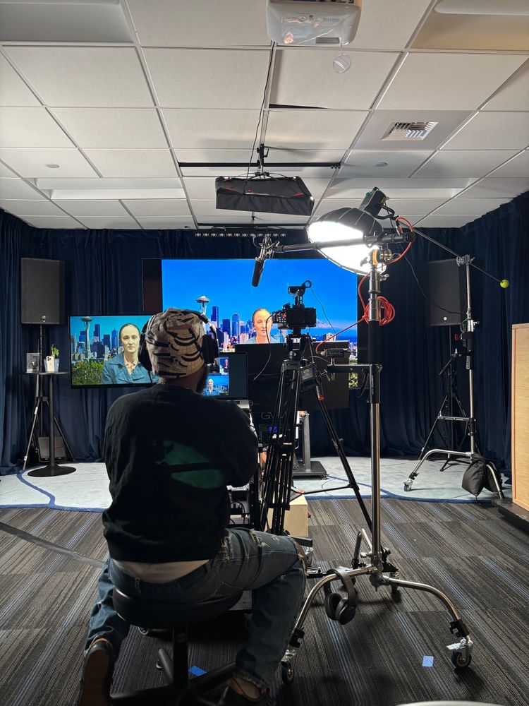 A man sits in a beanie with studio equipment in front of him and Katie Wilson sits in front of the cameras.