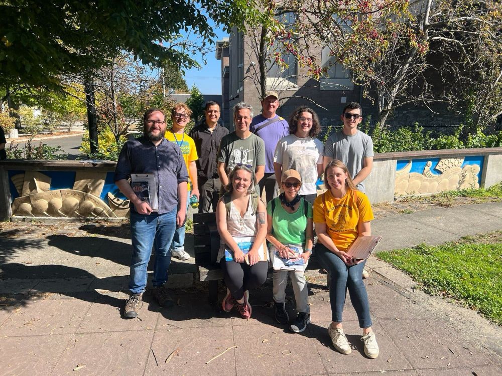 A group of 10 people hold clipboards and campaign literature before a canvas on a sunny day in a park.