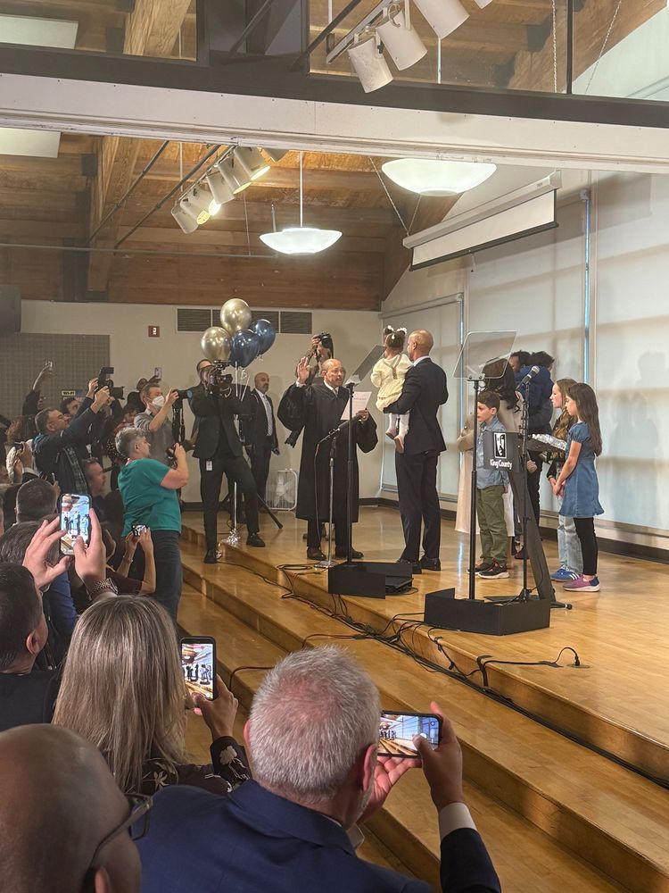 A photo of incoming king county executive girmay zahilay being sworn in on stage while holding a child, presumably his own, with other children on stage along with balloons and several on workers and press taking photos of the moment