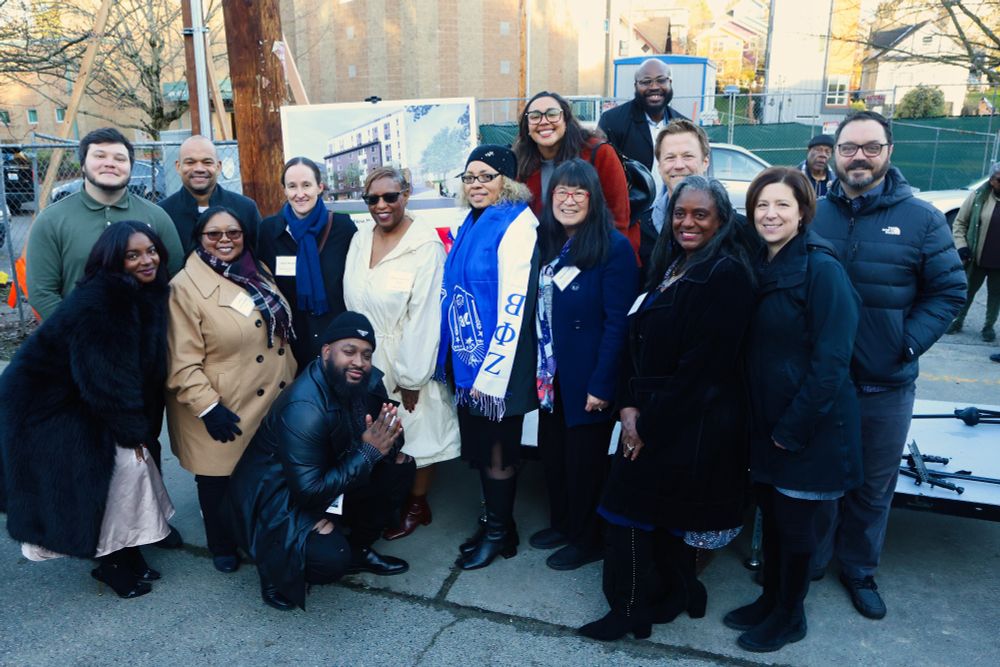 A photo of the group of community members and Katie Wilson posing for a group photo outside with a picture of the housing development behind them