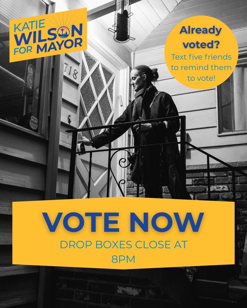 In a black and white image, Katie Wilson slips a piece of campaign literature into a doorknob of a home. She wears a thigh-length coat and there is a porchlight on behind her. The graphics on the image are blue text with yellow background and say, “vote now, drop boxes close at 8pm”; “Katie Wilson for Seattle Mayor;” and “already voted? Text five friends to remind them to vote!”