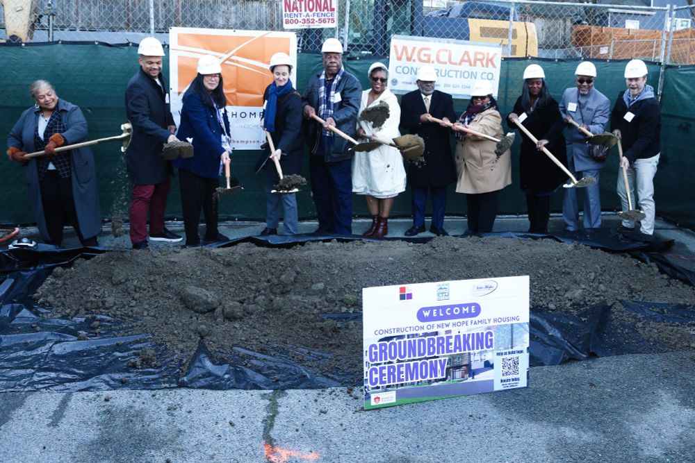 A photo of Katie Wilson and community members standing in a line before a patch of open dirt. they are wearing white hard hats, winter coats and holding shovels each with a scoop of dirt