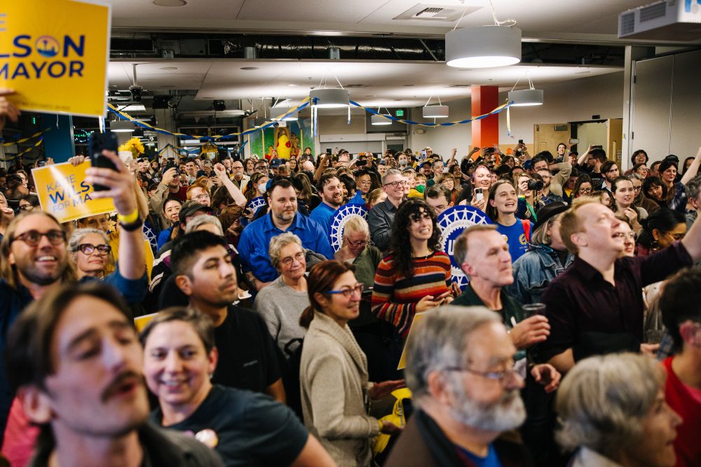 A photo of the packed crowd at the Katie Wilson election night party at El Centro de La Raza. supporters stand with signs and with arms raised