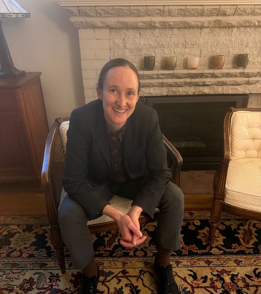 A photo of Katie Wilson seated in a wooden chair beside a white brick fireplace smiling at the camera with her hands folded