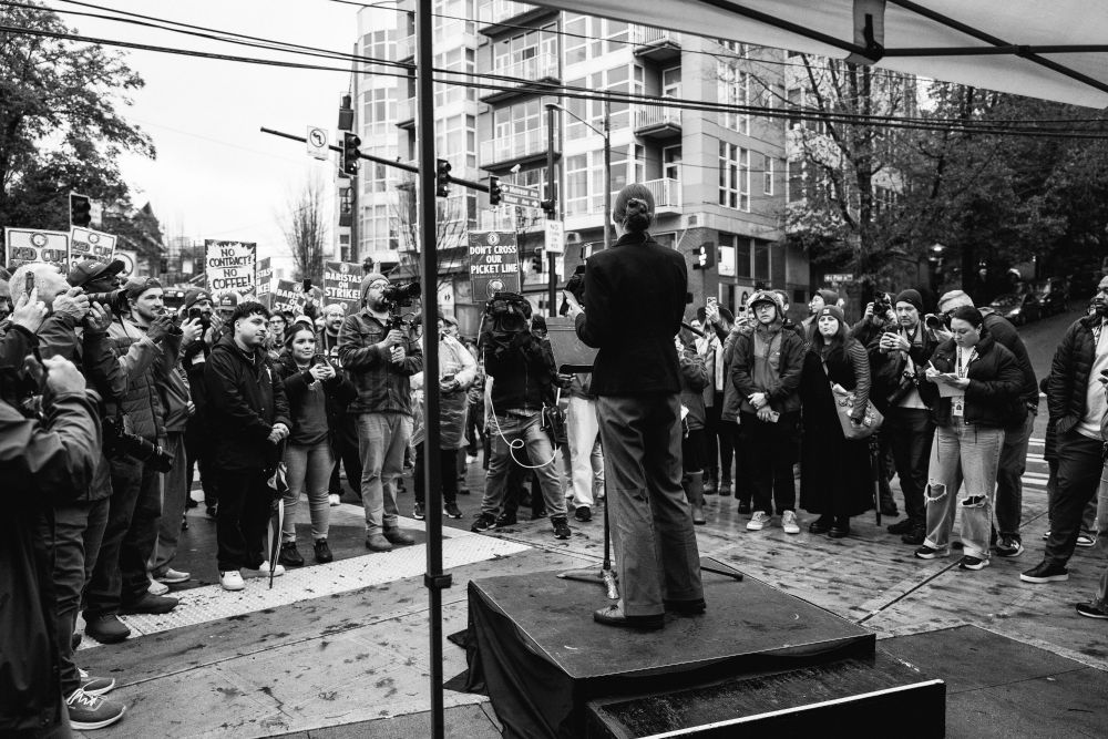 A black and white photo of Katie Wilson on a small stage speaking to a large crowd of people at the Starbucks workers rally