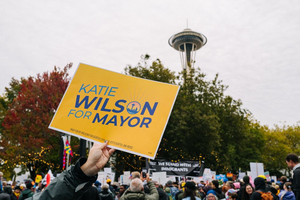 A color photo of a Katie Wilson for mayor sign held aloft above the crowd with the Space Needle in the background.