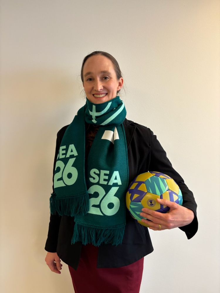 a photo of Mayor elect Katie Wilson standing before a white wall wearing the official world soccer Seattle 2026 scarf and holding a brightly colored soccer ball