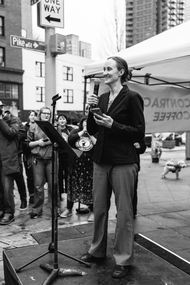 A black and white photo of Katie Wilson. standing with a microphone on a small stage outside on the sidewalk of Pike Street. a small crowd of onlookers are on her right