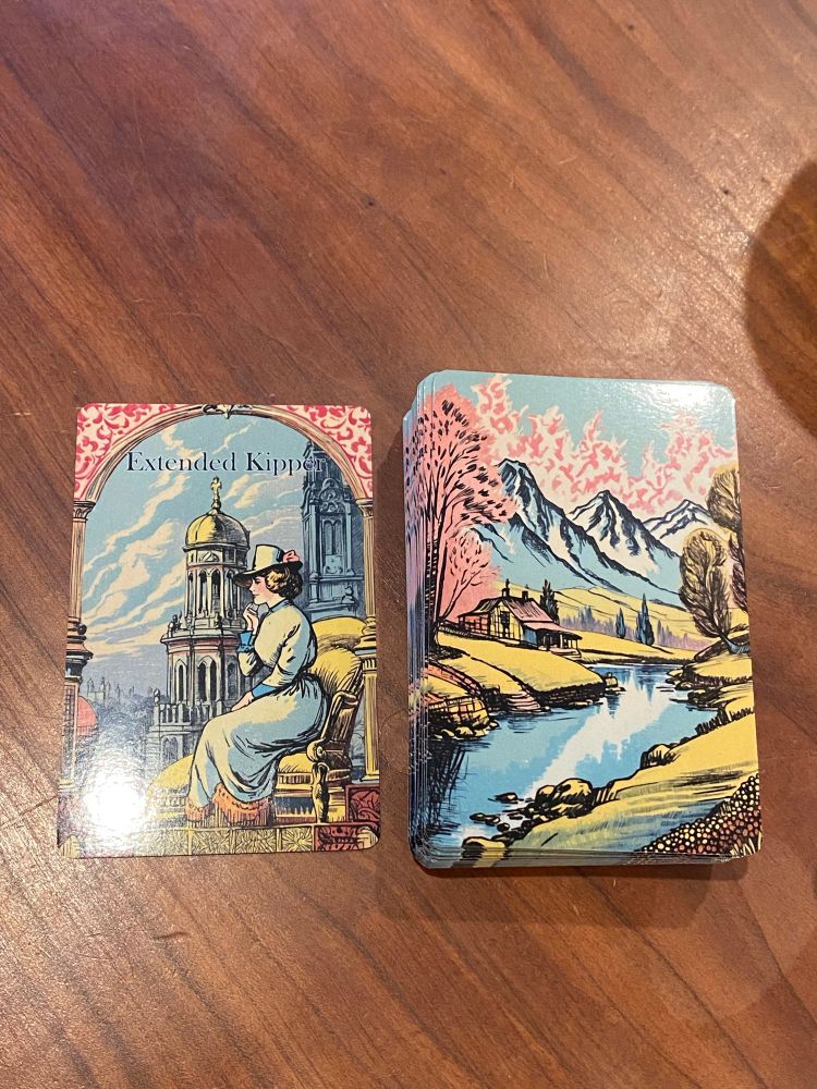 A deck of cards sit on a dark wood table. 

One card sits to the left of the deck, which is facedown, and reads, “Extended Kipper,” with a woman in Victorian attire sitting in a yellow chair with a tall tower in the background. 

The back of the deck shows a Bavarian Alps scene with a small cottage beside a river in a valley with flowering trees with pink blossoms and the snowcapped Alps in the background.

The deck is by Alex Bruinsma, available in their Etsy shop, OracleCards2023