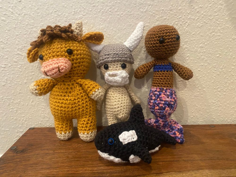 Four crocheted amigurumi are arranged on a brown wooden chest against an off-white wall. From left to right they are a: highland cow, a Viking, a bald mermaid, and in the front center is an orca. 