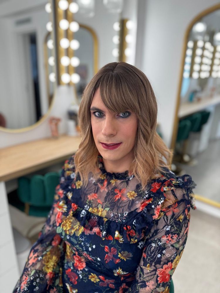 The image shows a person seated in a well-lit, stylish dressing room or beauty salon. The background features large mirrors framed with bright Hollywood-style lights and a sleek, modern decor with green velvet chairs.

The person has shoulder-length wavy hair in a soft brown tone with blonde highlights and a side-swept fringe. Their makeup is elegantly done, with defined eyeliner, dark red lipstick, and a soft blush. They’re wearing a beautiful dark navy dress with a sheer floral overlay in vibrant colors like red, yellow, and green. The overall look is feminine, polished, and confident, with a vintage-meets-modern vibe.