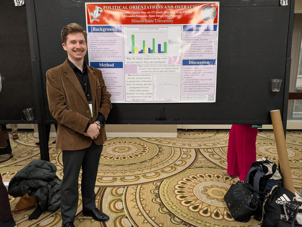 Aidan in a brown coat stands in front of a poster