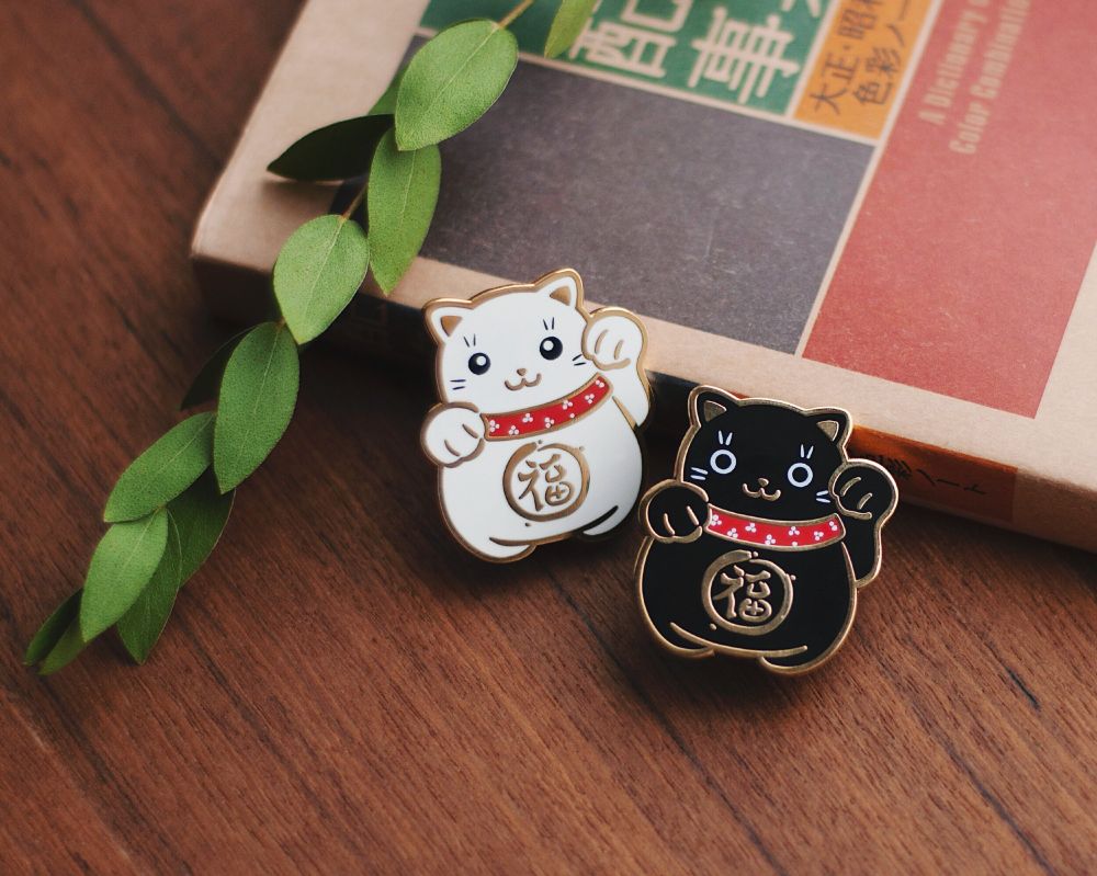 Two enamel pins depicting the Japanese lucky cat—one in black, one in white with gold trim.